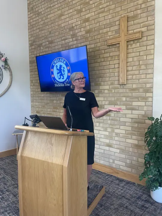 CLAIRE EVANS-BELL: Celebrant speaking at a wooden lectern beside a wall-mounted screen showing a football club crest