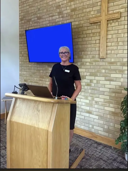CLAIRE EVANS-BELL: Smiling woman at a wooden lectern with laptop in a brick-walled room with a wooden cross and a blue screen.